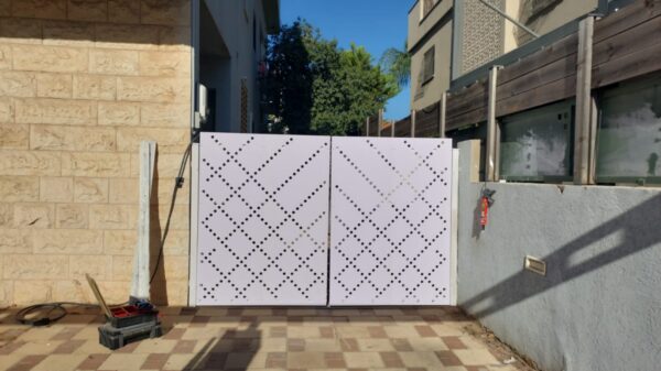 תמונה של שער כניסה לבן עם עיצוב מודרני, המורכב משתי דלתות משרביות מחוררות בדוגמת ריבועים ונקודות. משמאל לשער נראה קיר מחופה אבן בגווני חום בהיר, ומימין קיר בטון אפור. ברקע נראים עצים ומבנים נוספים, ורצפת השביל המוביל לשער מרוצפת באריחים בגווני חום בהיר.